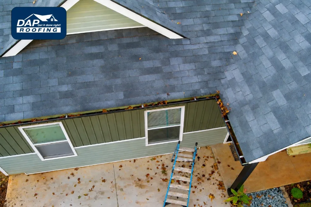Aerial view of a gray asphalt shingle roof with autumn leaves in the gutters and a ladder below